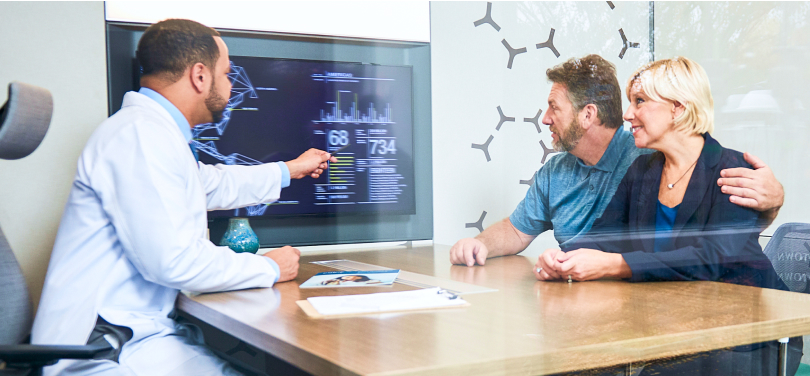 Man and woman sitting at a table with a healthcare professional looking at a TV screen displaying health data