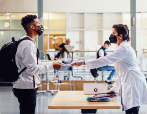 Male student wearing a mask and backpack receiving a Color test kit from a woman wearing a lab coat