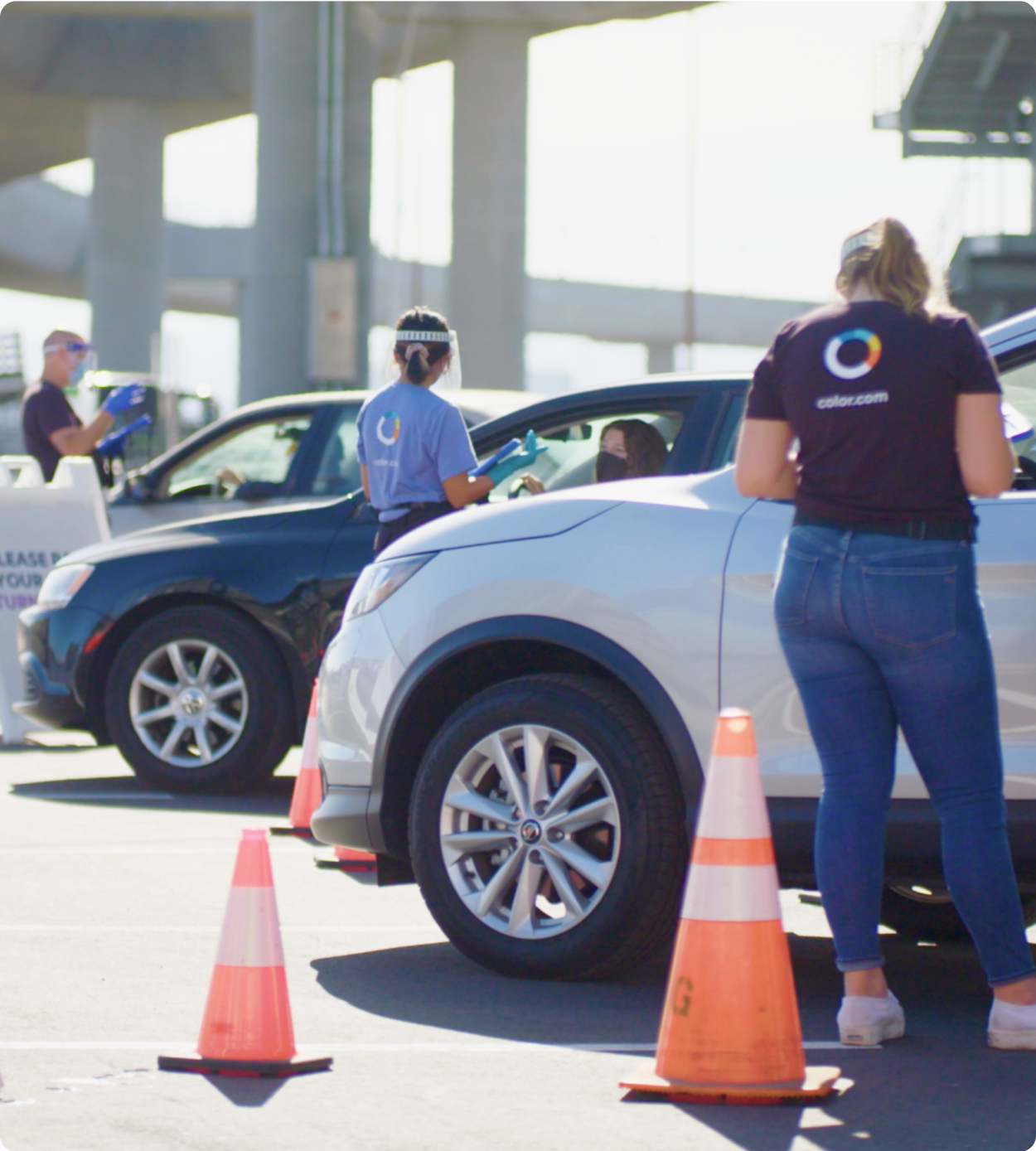 People wearing Color shirts standing next to cars and cones at a COVID-19 testing site