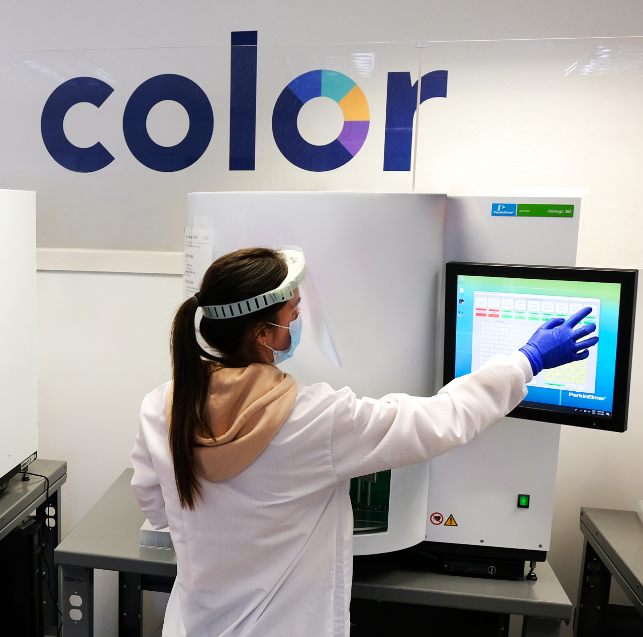 Woman wearing a lab coat and face shield pressing a screen while standing under a Color sign on the wall