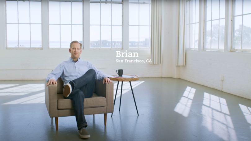 Man sitting in a chair in an empty room surrounded by windows with a cup of next to him