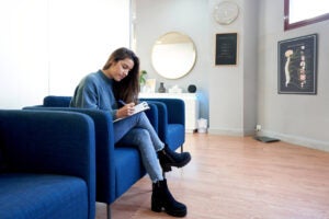 One girl in casual wear sitting in waiting room of hospital and filling in form, medical poster on wall. Young female visiting doctor. Concept of healthcare