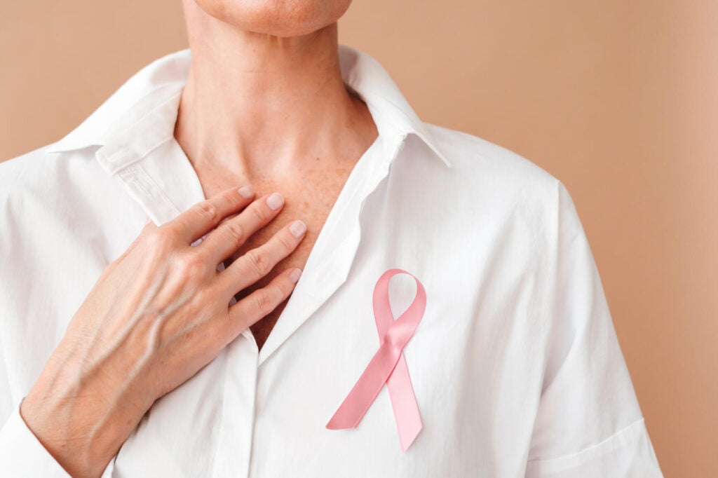 Anonymous woman wearer in white shirt with pink ribbon as symbol of Awareness and fight against breast cancer keeps hand on chest. Breast Cancer Awareness Day concept.