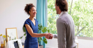 Young black woman in blue dress shaking hands with young white man wearing a gray knit sweater in an office setting.
